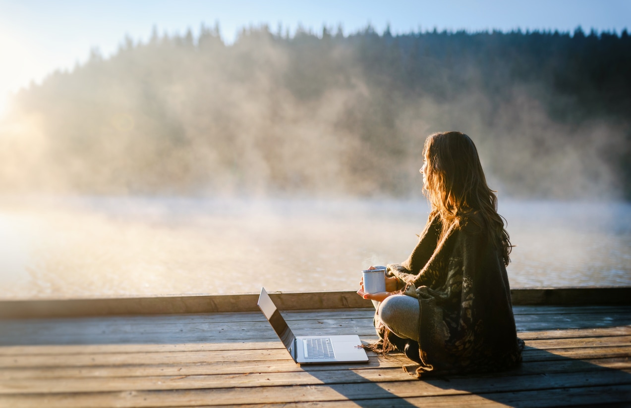 Frau Remote Work am See Eine Frau sitzt mit einer Tasse in der Hand und ihrem Laptop an einem See und arbeitet Remote
