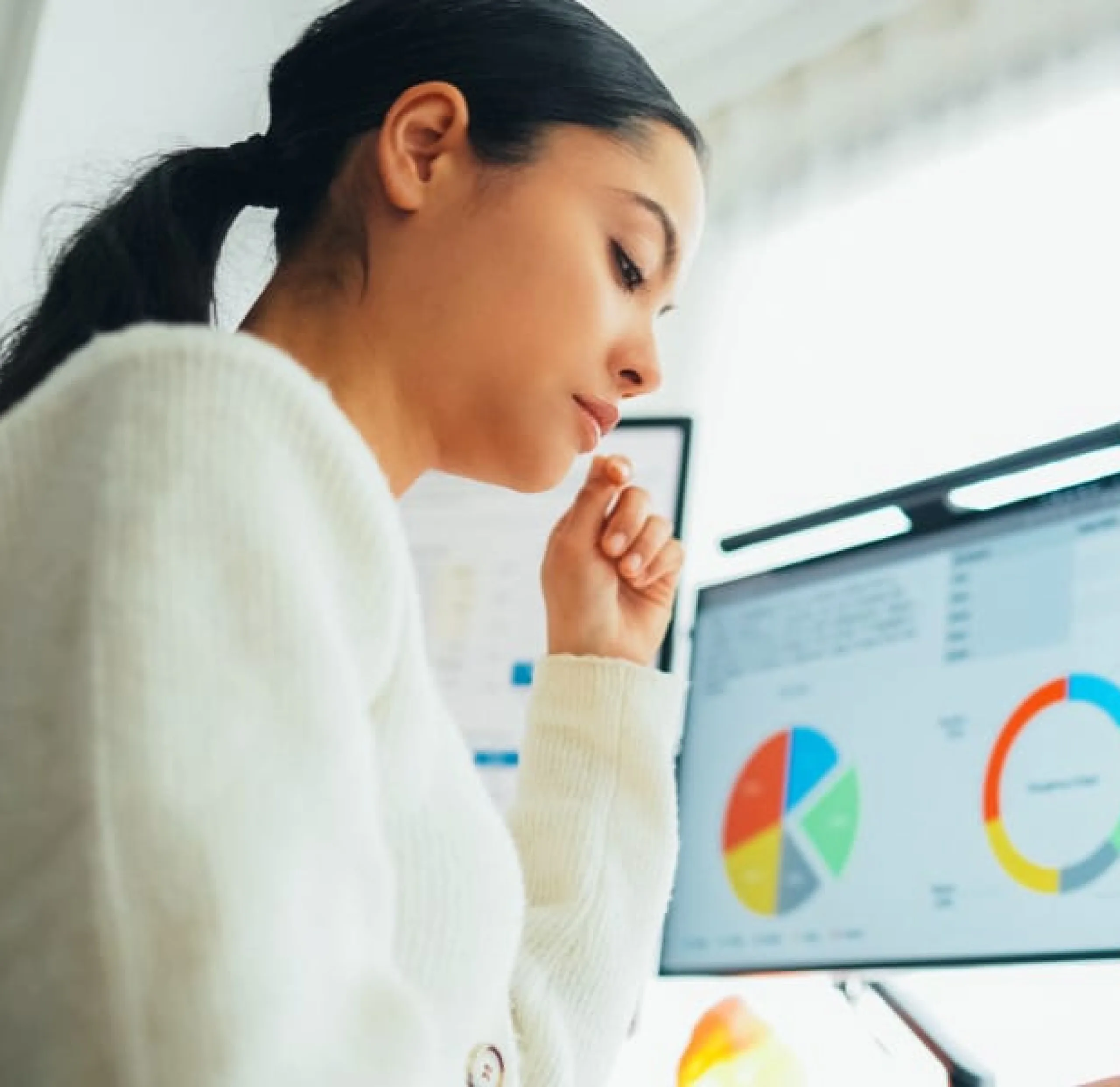 Eine junge Frau mit dunklem Haar und weißem Pullover sitzt konzentriert vor einem Computerbildschirm. Auf dem Bildschirm sind verschiedene Diagramme und Grafiken zu sehen. Das Bild veranschaulicht die Digitalisierung im Vertrieb.