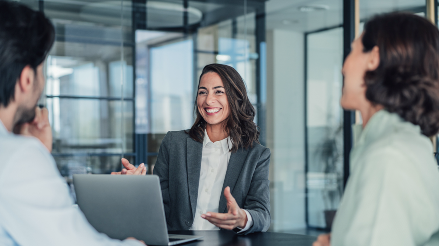 business-gross Eine lachende Geschäftsfrau in einem grauen Blazer sitzt an einem Konferenztisch und spricht mit zwei Kollegen. Vor ihr steht ein aufgeklappter Laptop, und die Umgebung scheint ein modernes Büro mit Glaswänden zu sein.