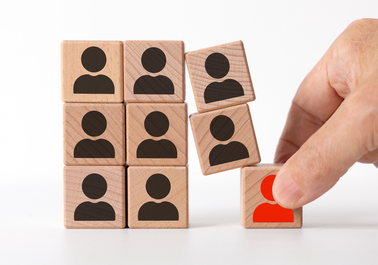 personalentscheidung-symbolbild Eine Hand greift nach einem Holzbaustein mit einem roten Symbol einer Person, der sich von anderen Bausteinen mit schwarzen Symbolen abhebt, die in einem 3x3-Gitter angeordnet sind. Das Bild symbolisiert eine Personalentscheidung, bei der eine bestimmte Person ausgewählt oder entfernt wird.
